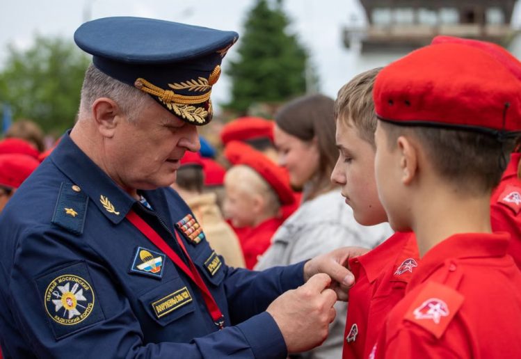 A Russian officer in uniform pinning a badge on a young recruit during an outdoor military ceremony.