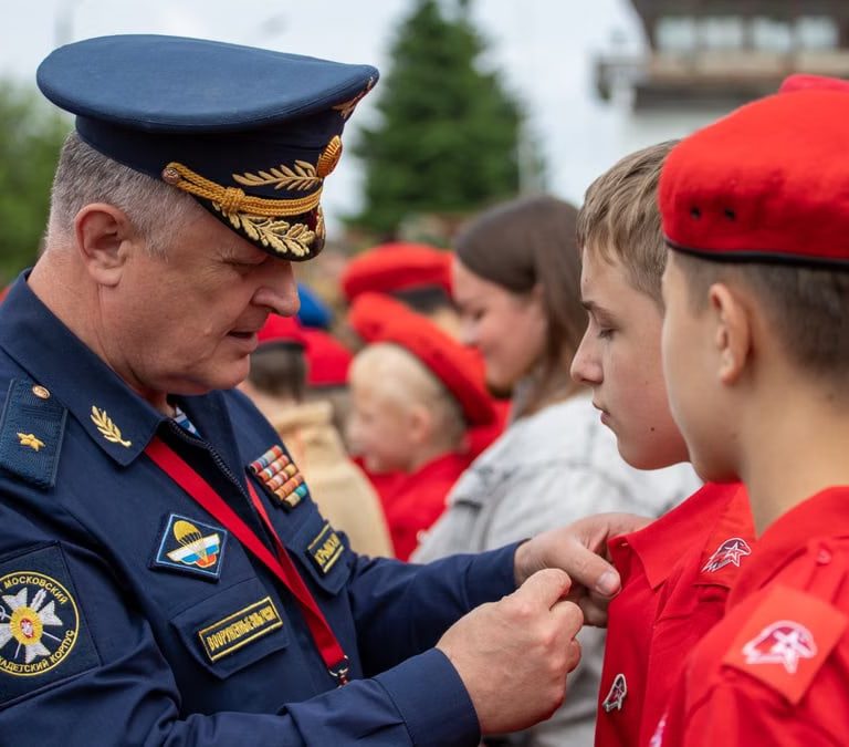 A Russian officer in uniform pinning a badge on a young recruit during an outdoor military ceremony.