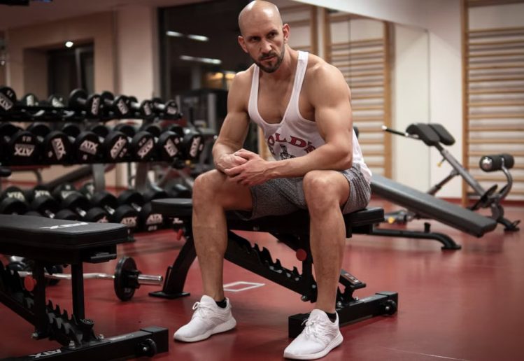 Strong, determined man in white tank top sitting in gym, showcasing fitness and strength.