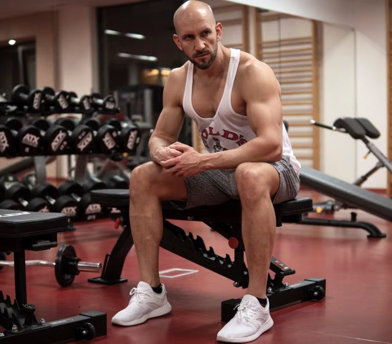 Strong, determined man in white tank top sitting in gym, showcasing fitness and strength.